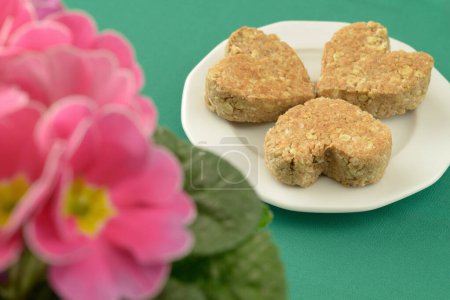 Photo for Heart-shaped cookies on a white plate. Pink Primroses in the foreground. Cookies made with organic oatmeal, coconut flakes and wholemeal flour. - Royalty Free Image