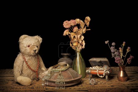 Osito de peluche vintage de los años 50 sentado cerca de objetos de aspecto retro como un pequeño cofre lleno de joyas, jarrones con flores marchitas y otros artículos de decoración. Fondo negro.