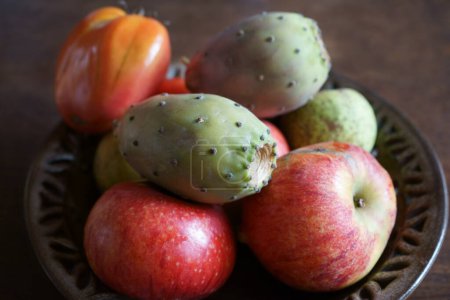Ein brauner Teller auf einem braunen Holztisch, gefüllt mit Früchten wie Kaktusfeigen, Äpfeln, Birnen und Tomaten.