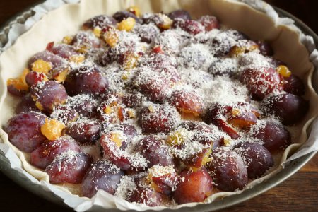 A raw plum pie with coconut flakes in a round cake pan. Ready for the oven. Close-up.
