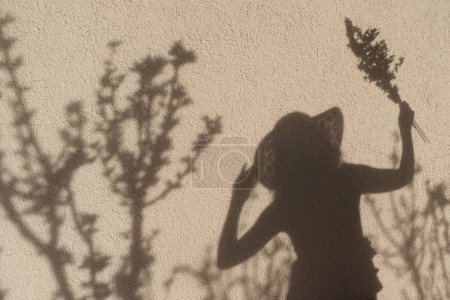 Schatten einer Frau an einer Wand. Sie trägt einen Strohhut und hält Blumen in der Hand. Schatten von Pflanzen um sie herum. 