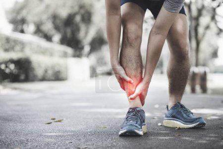 Concepto de lesión física: Hombre asiático sosteniéndose el tobillo mientras corre por la calle en el parque.