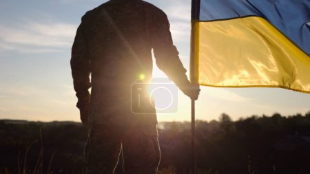 Ein ukrainischer Armeeangehöriger steht bei Sonnenuntergang mit einer Nationalfahne auf dem Land. Männlicher Soldat in Militäruniform mit ukrainischer Flagge als Symbol des Sieges über die russische Aggression. Invasionswiderstandskonzept