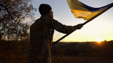 Young man in military uniform waving flag of Ukraine against beautiful sunset at background. Male ukrainian army soldier lifted national banner at countryside. Victory against russian aggression.