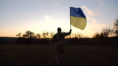 Soldat der ukrainischen Armee läuft mit erhobenem blau-gelbem Banner in der Abenddämmerung über das Feld. Junge Männer in Uniform joggen mit der Fahne der Ukraine auf der Wiese. Sieg gegen russisches Aggressionskonzept.