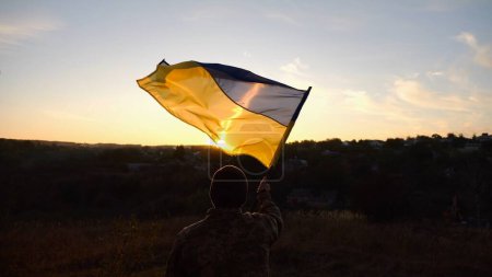 Jeune homme en uniforme militaire agitant le drapeau de l'Ukraine contre un beau coucher de soleil en arrière-plan. Soldat ukrainien masculin levé bannière nationale à la campagne. Victoire contre l'agression russe.