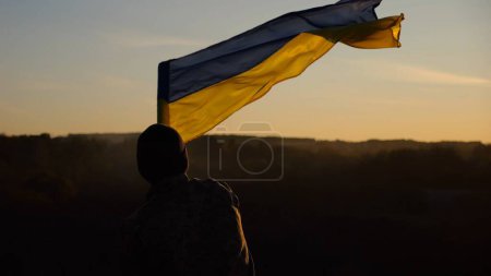 Junger Mann in Militäruniform schwenkt die Flagge der Ukraine vor dem schönen Sonnenuntergang im Hintergrund. Männlicher ukrainischer Armeesoldat hisste auf dem Land die Nationalfahne. Sieg gegen die russische Aggression.