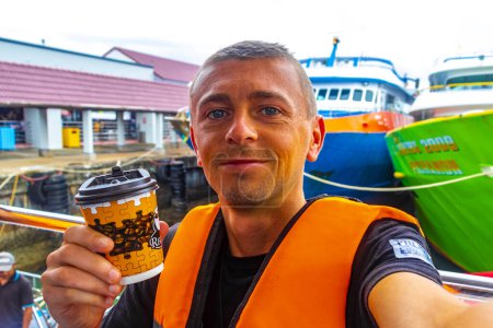 Téléchargez les photos : Homme beau touriste avec café et gilet de sauvetage ouest sur un ferry pour Ao Nang à Ratsada Amphoe Mueang Phuket sur l'île de Phuket Thaïlande en Asie du Sud-Est. - en image libre de droit