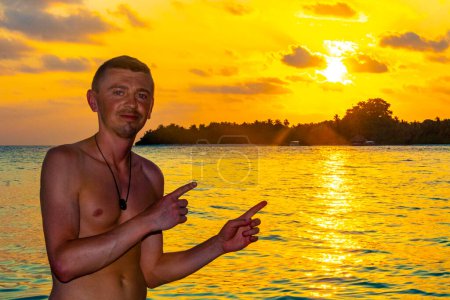 Foto de Joven guapo turista masculino en la isla del paraíso tropical Kuramathi para unas vacaciones de lujo con palmeras de arena blanca aguas turquesas y una increíble puesta de sol de colores dorados en el atolón Rasdhoo Maldivas. - Imagen libre de derechos