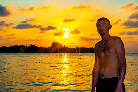 Foto de Joven guapo turista masculino en la isla del paraíso tropical Kuramathi para unas vacaciones de lujo con palmeras de arena blanca aguas turquesas y una increíble puesta de sol de colores dorados en el atolón Rasdhoo Maldivas. - Imagen libre de derechos