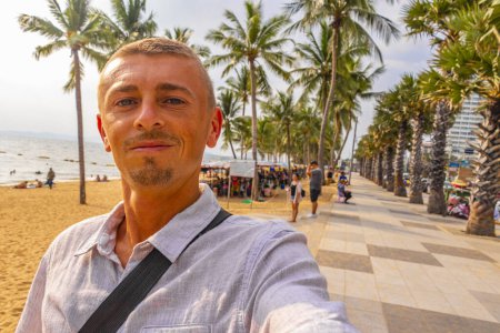 Tourist man takes selfie photo at tropical beach waves water sand people boat palm trees and skyscrapers in Jomtien Beach Pattaya Bang Lamung Chon Buri Thailand in Southeastasia Asia.