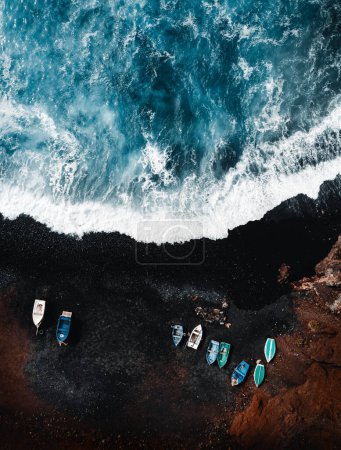 Photo for Vertical aerial photo of black sand volcanic beach with boats on the shore and big and strong waves in ocean -  Lanzarote, Canary Islands. Waves in blue ocean from above (top view) with red cliff. - Royalty Free Image