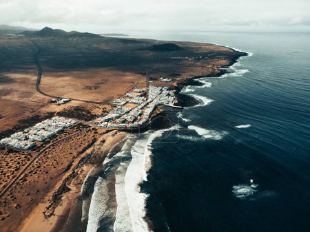 Photo for Aerial photo of sunset on Caleta de Famara in Canary Islands - Lanzarote:  wide photo from above with strong waves in blue ocean and white clear sky. Cliffs and rocks with ocean by drone. - Royalty Free Image