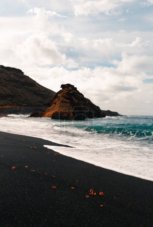Photo for Vertical photo of black sand El Golfo beach in volcanic island Lanzarote (Canary Islands) with strong waves in blue ocean. Cliff in wild black sand beach with  cloudy blue sky on background. - Royalty Free Image