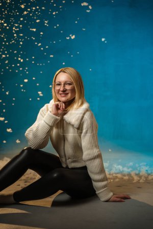 Photo for Blonde cute caucasian woman in white fur sweater on blue background. Fake Snow is flowing all over her. She is happy, celebrating christmas and new year. Studio portrait - Royalty Free Image