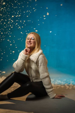Téléchargez les photos : Blonde mignonne femme caucasienne en pull en fourrure blanche sur fond bleu. Fausse neige coule sur elle. Elle est heureuse, fêtant Noël et le nouvel an. Portrait studio - en image libre de droit