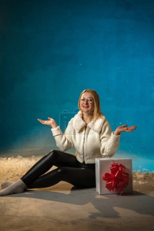 Téléchargez les photos : Blonde mignonne femme caucasienne en pull en fourrure blanche sur fond bleu. Fausse neige coule sur elle. Elle est heureuse, assise avec un cadeau, célébrant Noël et le nouvel an. Portrait studio - en image libre de droit