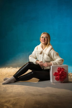 Téléchargez les photos : Blonde mignonne femme caucasienne en pull en fourrure blanche sur fond bleu. Fausse neige coule sur elle. Elle est heureuse, assise avec un cadeau, célébrant Noël et le nouvel an. Portrait studio - en image libre de droit