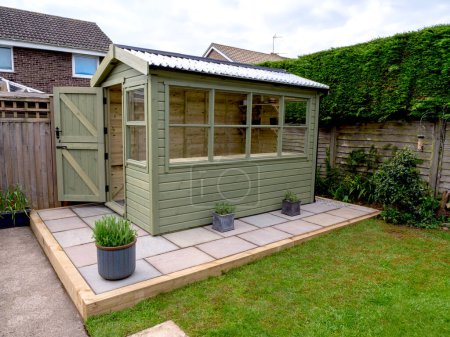 Verde cobertizo de madera manchada con ventanas de cristal y una puerta abierta en un patio pavimentado en un jardín