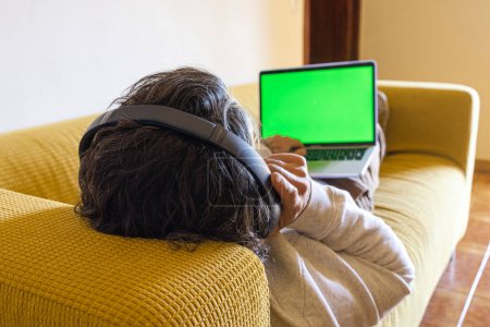 Téléchargez les photos : Homme détendu assis sur le canapé à la maison avec écouteurs et ordinateur portable - en image libre de droit