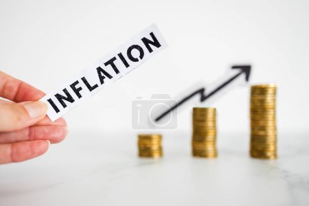 Hand hält Inflationstext vor wachsenden Stapeln von Münzen mit Pfeil nach oben im Hintergrund aufgenommen in geringer Schärfentiefe