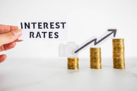 Photo for Hand holding Interest Rates text in front of growing stacks of coins with arrow going up  in the background, symbol of excessive pricing or speculation - Royalty Free Image