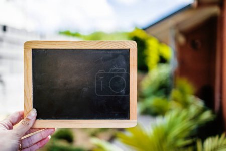 Photo for Hand holding small blackboard with blank copy space in front of stylish backyard and house exterior background, space to add your text - Royalty Free Image