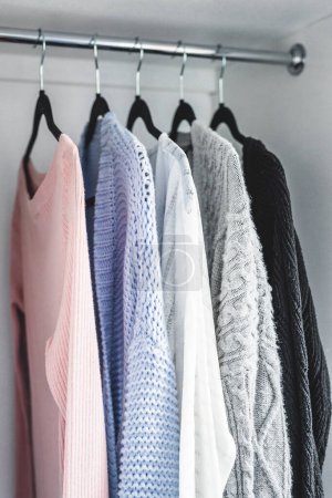 minimalist capsule wardrobe selection of womenswear sweaters and cardigans hang on wardrobe rod with matching hangers shot at shallow depth of field