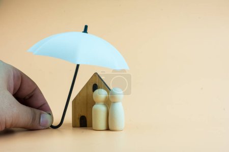 Foto de Family of wooden dolls and home are under a white umbrella, protecting wooden peg dolls and home, planning, saving families, preventing risks and crises, and insurance concepts. - Imagen libre de derechos