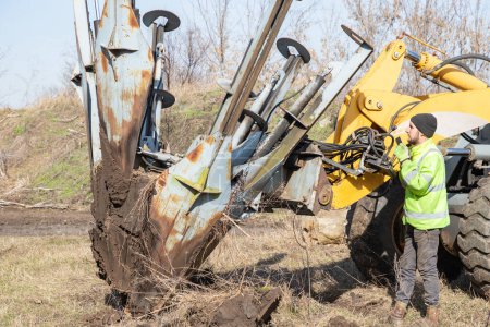 Male worker operates tree transplanter heavy machine. Landscaping, seasonal agricultural engineering, large trees landing machines. Planting of tree using tree spade - specialized machine for transplanting and transport trees.