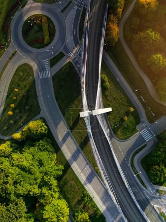 Luftaufnahme direkt von Straßenring und Hängebrücke in Krakau, Polen. Malerisches Sonnenlicht und Schatten auf den Baumwipfeln des Stadtparks, leere Straße ohne Autos und moderne Straßenbahnbrücke