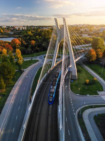 Drone forward overhead view of suspension bridge with tram movement in Krakow, Poland. Sunset top view of bridge structure and public transport in motion blur, empty asphalt road and park below