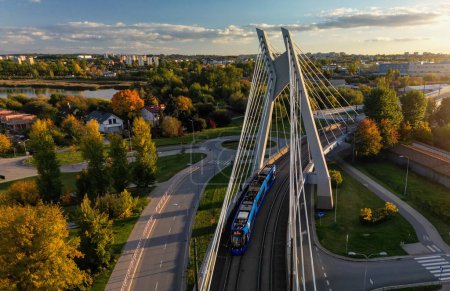 Aerial perspective view of city road and modern bridge in Krakow, Poland. Drone forward above view of tram driving on circular steel and concrete bridge construction, autumn park in sunset rays