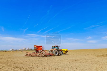 Foto de Agricultural machinery works during spring field work. A tractor with a seeder and an additional container for sowing seeds is sowing grain in the field. Agro-industrial business. - Imagen libre de derechos