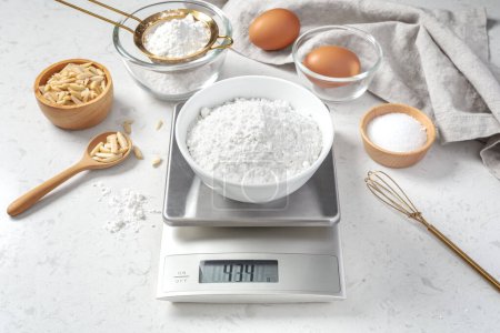 Foto de Harina en cuenco blanco que mide a escala digital con ingredientes para pasteles o panaderías y utensilios en mesa de cocina de mármol - Imagen libre de derechos