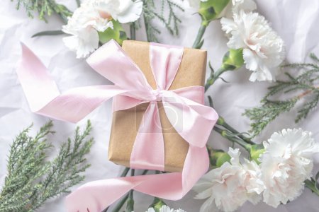 Photo for Gift box wrapped in craft paper with pink bow on natural flowers and green leaves background, top view - Royalty Free Image
