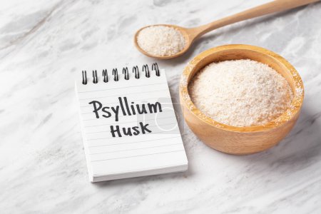 Cáscara de Psyllium en cuenco de madera y cuchara con la palabra de la cáscara de psyllium en el cuaderno en la mesa de mármol blanco