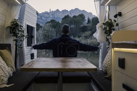 Hombre maduro de vacaciones abriendo las puertas traseras de una caravana estacionada frente a un hermoso paisaje de montaña. Vida de furgoneta conceptual, personas, viajes, transporte, limpieza