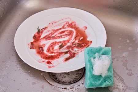 Foto de Una placa sucia y una esponja de espuma con líquido de lavado están en el fregadero de la cocina. - Imagen libre de derechos