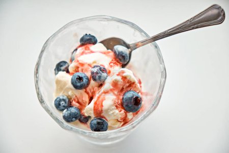 Foto de Placa de vidrio para crema llena de helado y arándanos rociados con jarabe de cereza con una cuchara de postre sobre un fondo blanco. - Imagen libre de derechos