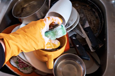 Foto de Un hombre con guantes protectores sostiene una esponja de cocina húmeda en su mano contra el fondo de platos sucios. - Imagen libre de derechos