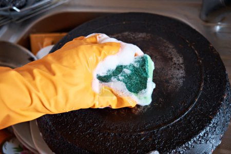 Una mano de hombre en un guante protector limpia una sartén con un fondo quemado con una esponja de cocina de espuma.