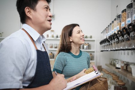 Photo for Asian male shopkeeper describes natural organic products to woman customer in refill store, zero-waste and plastic-free grocery, environment-friendly, sustainable lifestyles with reusable containers. - Royalty Free Image
