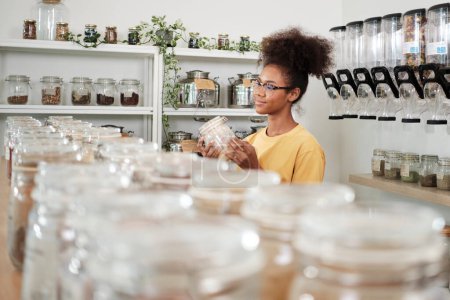 Photo for Young African American woman is choosing and shopping for organic products in refill store with reusable container, zero-waste grocery, and plastic-free, environment-friendly, sustainable lifestyles. - Royalty Free Image