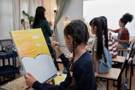 Eine Gruppe von multirassischen Kindern, die mit einer Asiatin lernen, unterrichtet Acrylfarbenmalerei auf Leinwand im Kunstunterricht, kreatives Lernen mit Geschick in der Atelierausbildung der Grundschule.