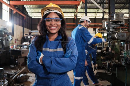 Téléchargez les photos : Portrait d'une travailleuse noire professionnelle en uniforme de sécurité et casque de protection regardant la caméra, les bras croisés et souriants, avec une équipe d'ingénieurs derrière elle dans une usine de fabrication de métaux. - en image libre de droit