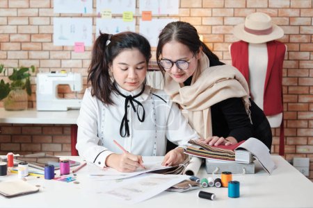 Téléchargez les photos : Asian middle-aged female fashion designer teaches a young teen trainee tailor in studio with colorful thread and sewing fabric for dress design collection ideas, professional boutique small business. - en image libre de droit