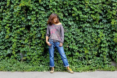 Photo for A young model beautiful woman girl in denim trousers stands near a wall overgrown with climbing plants. Background with plant leaves - Royalty Free Image