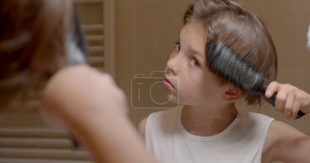 Photo for Boy combs his hair in the mirror, he holds a comb with his hand, makes a movement. Make hair style homemade haircut. - Royalty Free Image