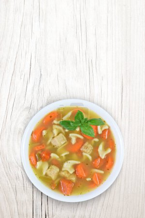 Top view of warm, home cooked chicken noodle soup with fresh carrots and extra chicken. Set atop a modern white wood background with plenty of copy space.
